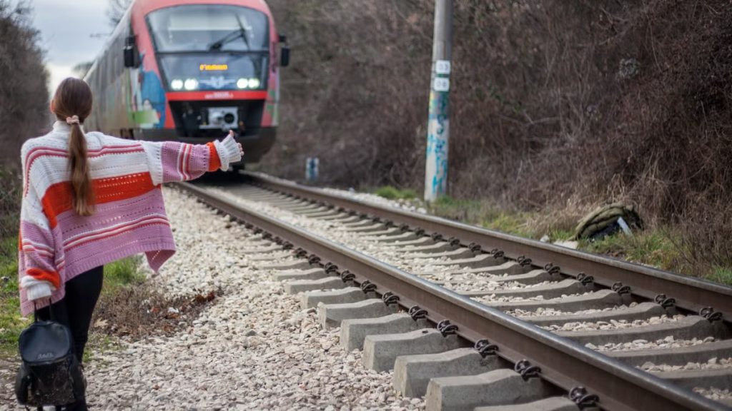 A woman in a colorful sweater hitchhikes near railway tracks as a train approaches in a rural setting.